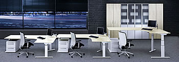 H&ouml;henverstellbarer Schreibtisch Spine&sup2;: Drei Eckschreibtische (240 x 160 x 80 cm) aufgestellt mit unterschiedlichen Arbeitsh&ouml;hen.Oberfl&auml;che in Ahorn-Dekor und kristallwei&szlig;es Gestell.